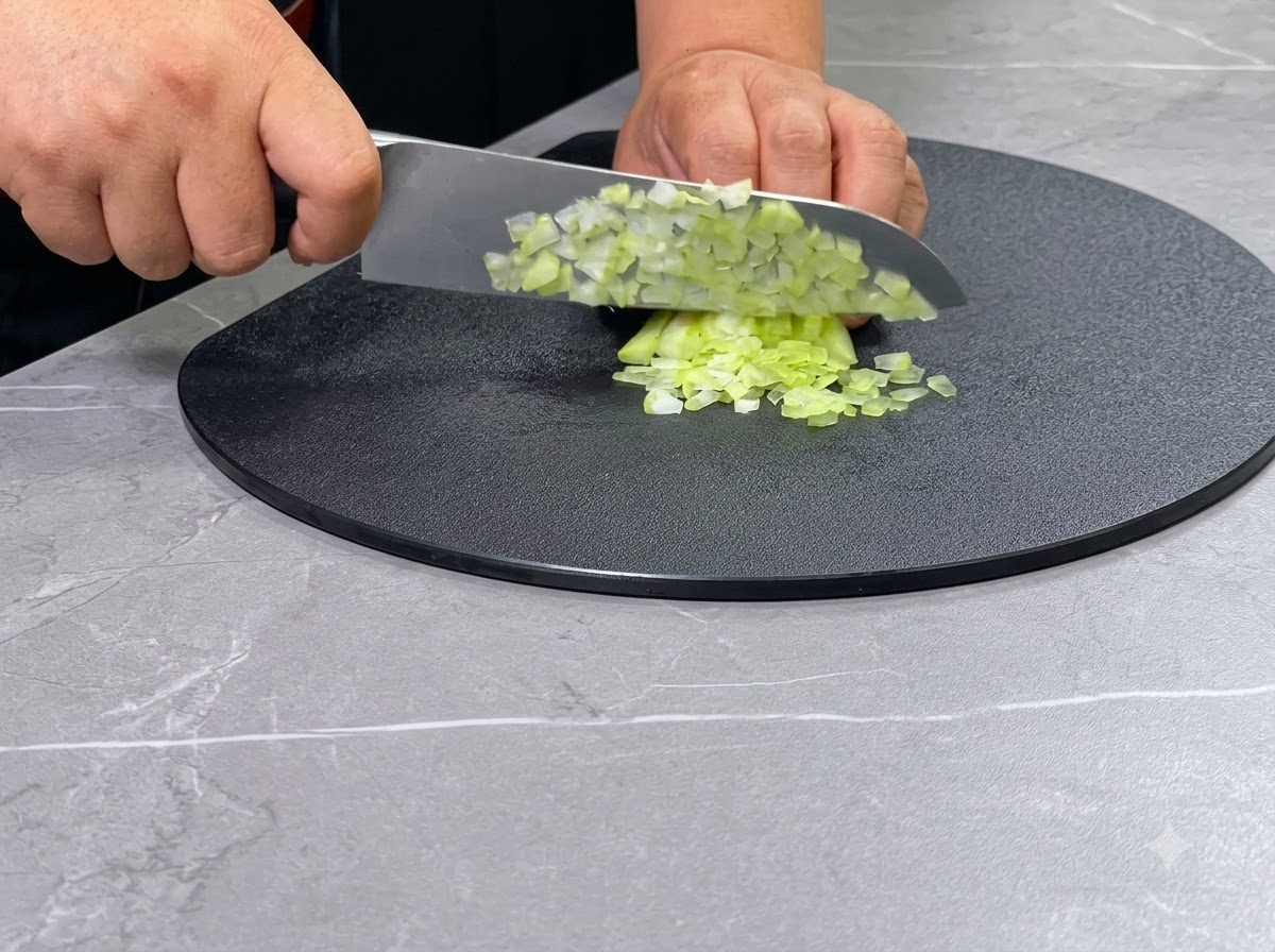 A pair of hands using a chefs knife to dice green celery stalks on a black circular cutting board.