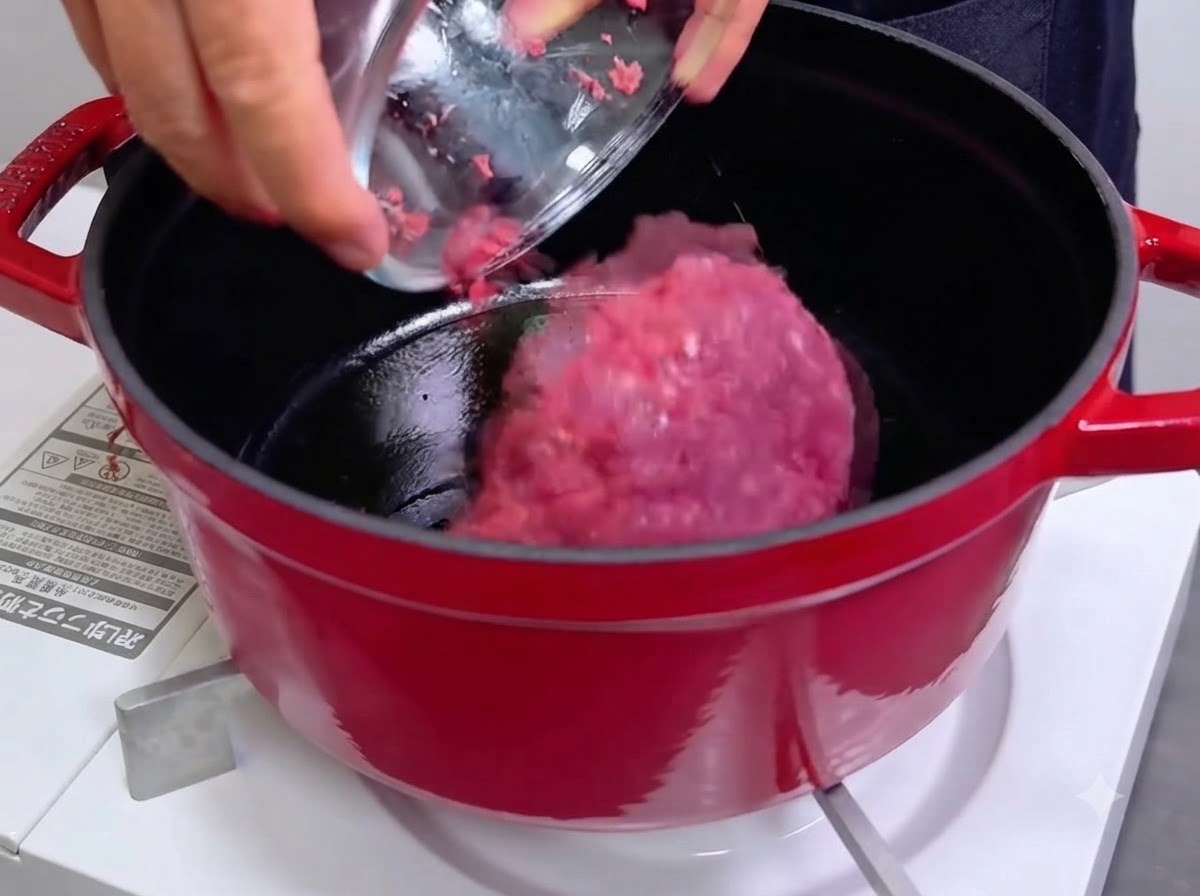 A bowl pouring pink minced raw meat into a hot red cast iron Dutch oven.