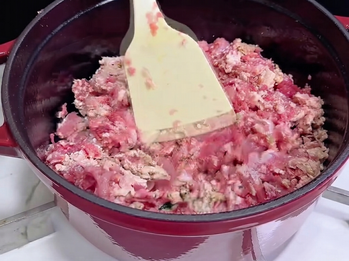 Using a white spatula to stir-fry minced meat in a red cast iron pot as it begins to brown.