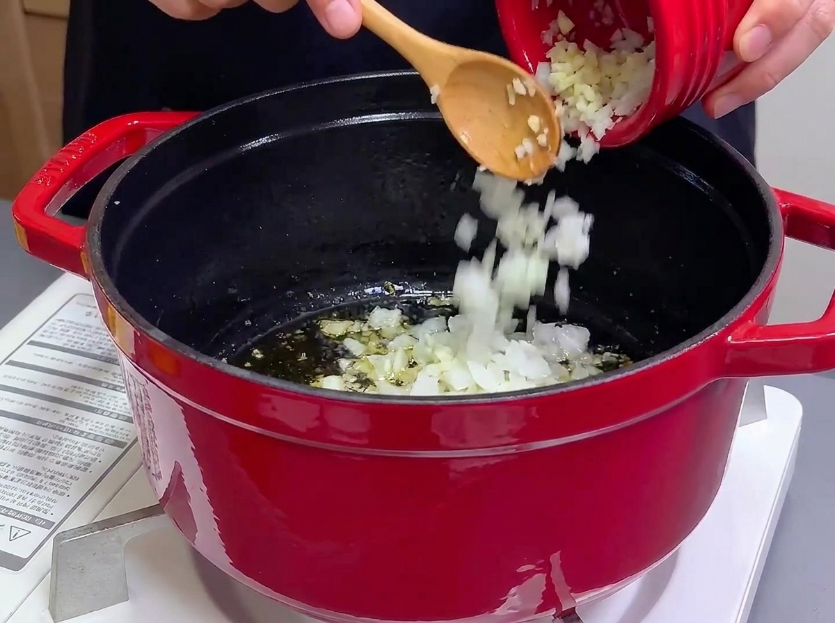 Pouring minced onions and garlic from a small red bowl into a cast iron pot with hot oil.