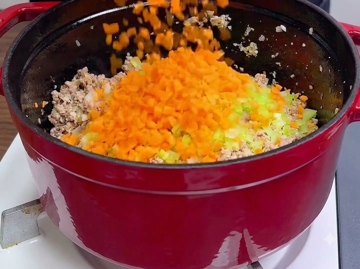 Freshly chopped orange carrots and green celery being poured directly over cooked minced meat inside a red pot.
