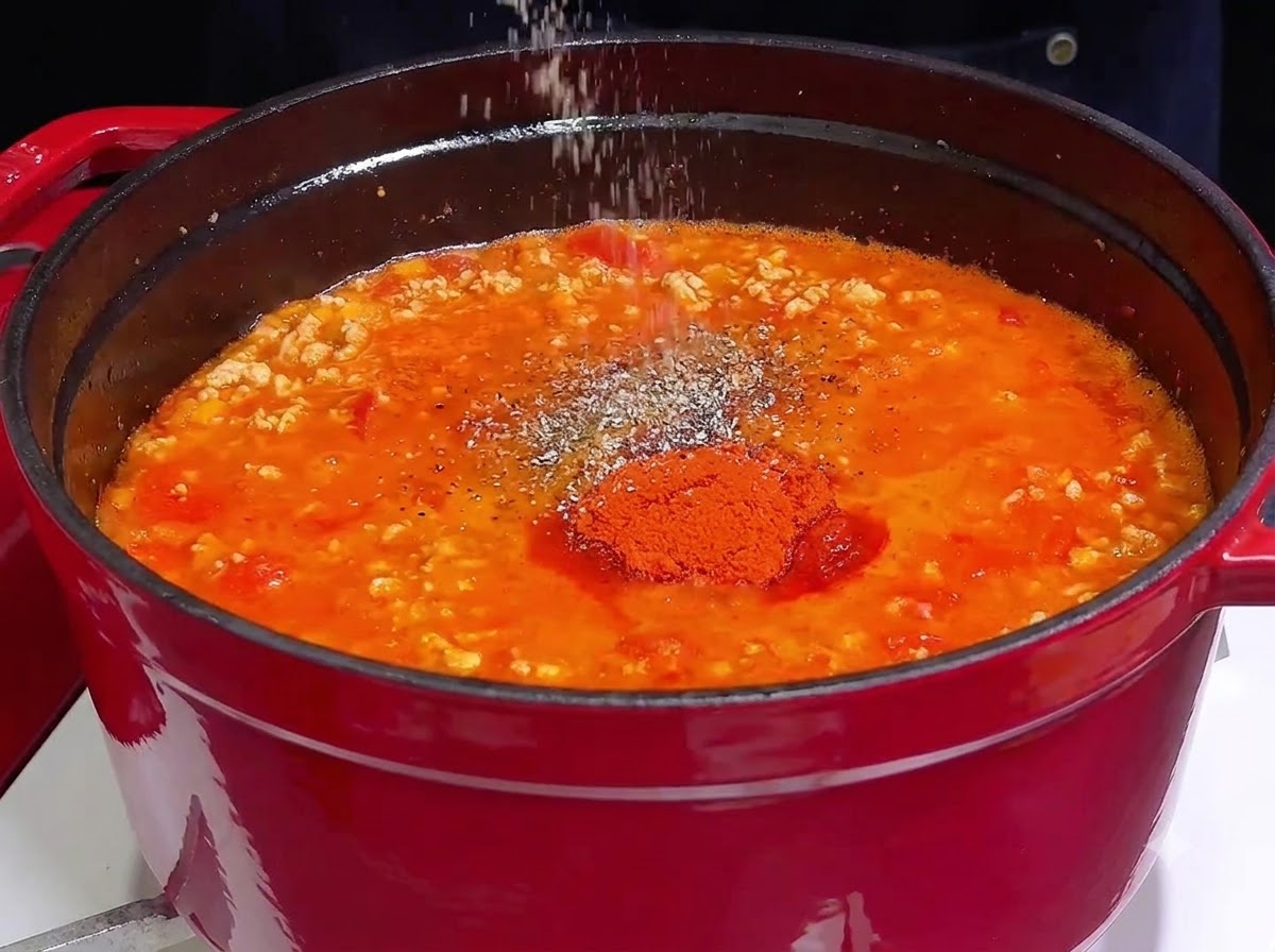 A hand sprinkling salt and spices over a vibrant red simmering meat sauce in a cast iron pot.