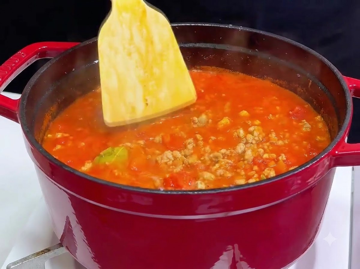 A wooden spatula stirring red wine into a red cast iron pot filled with bubbling tomato meat sauce.