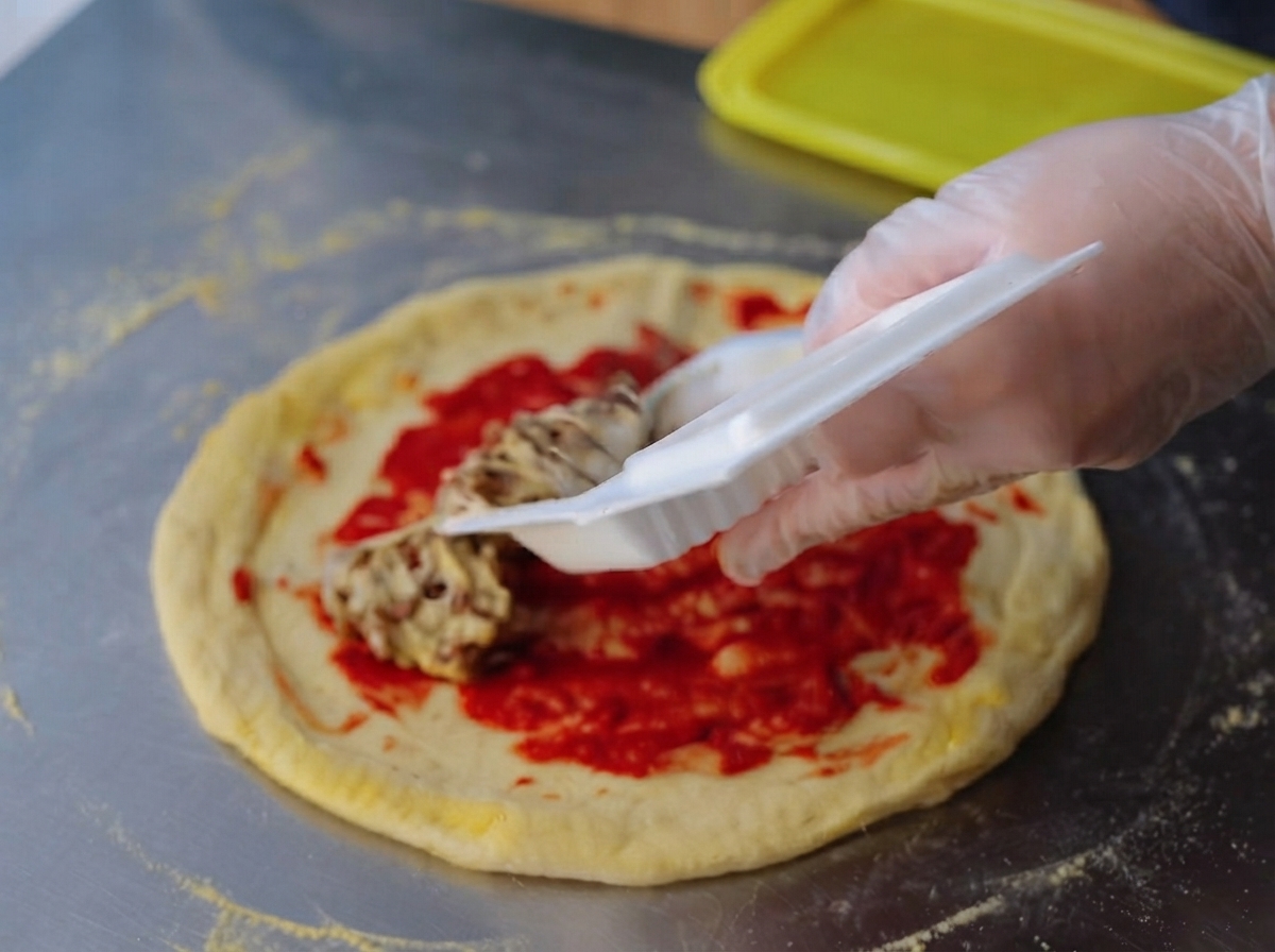 A gloved hand spreading a sticky, stringy natto mixture evenly over the bright red tomato paste layer on a raw pizza dough.