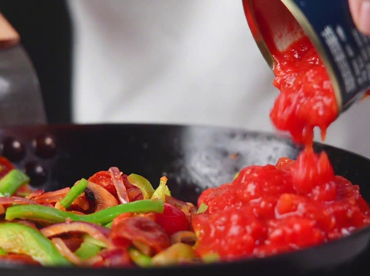 Canned chopped tomatoes being poured into the pan with vegetables and salami.