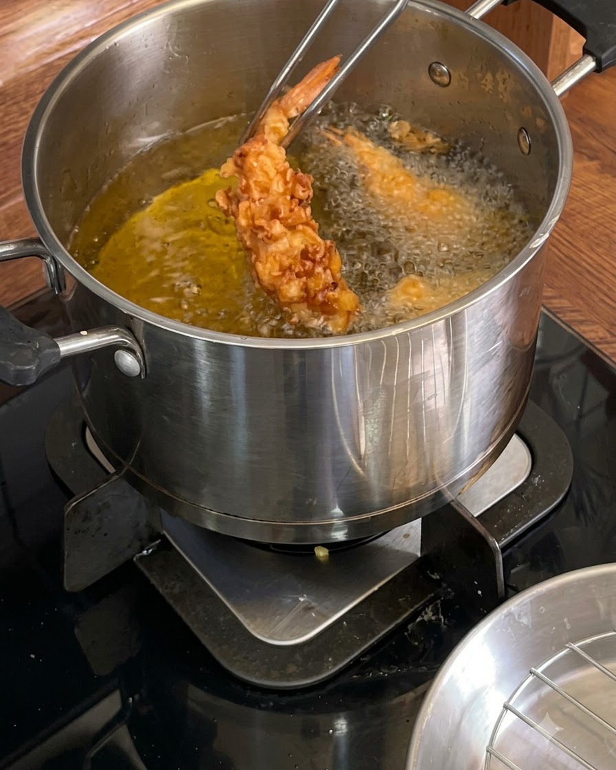 Metal tongs holding a golden-brown crispy fried shrimp over a hot pot of oil.