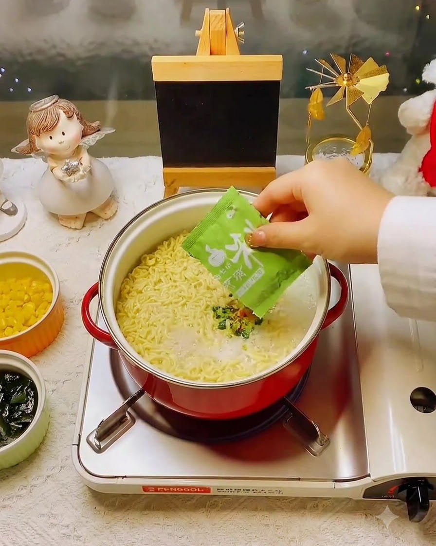 A hand adding a green seasoning packet into the pot of noodles and broth.