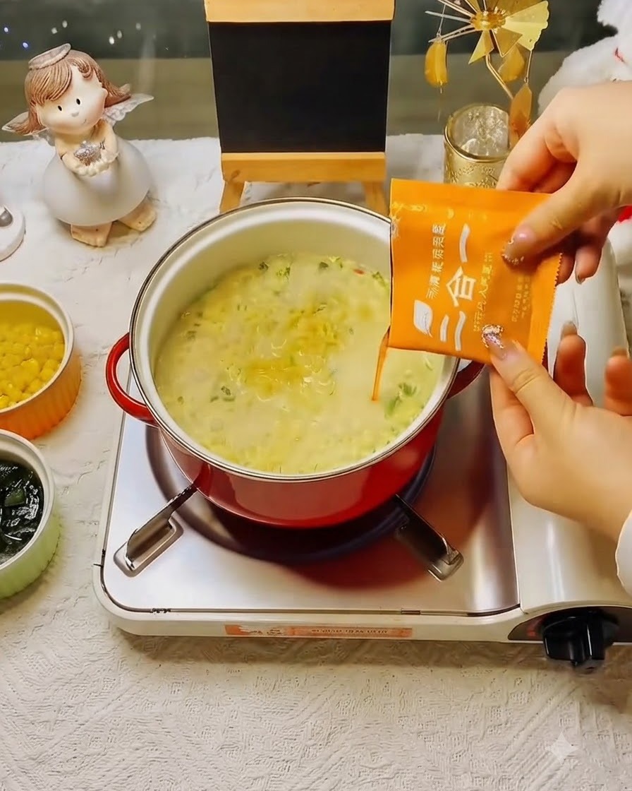 An orange seasoning soup packet being poured into the simmering pot of noodles.