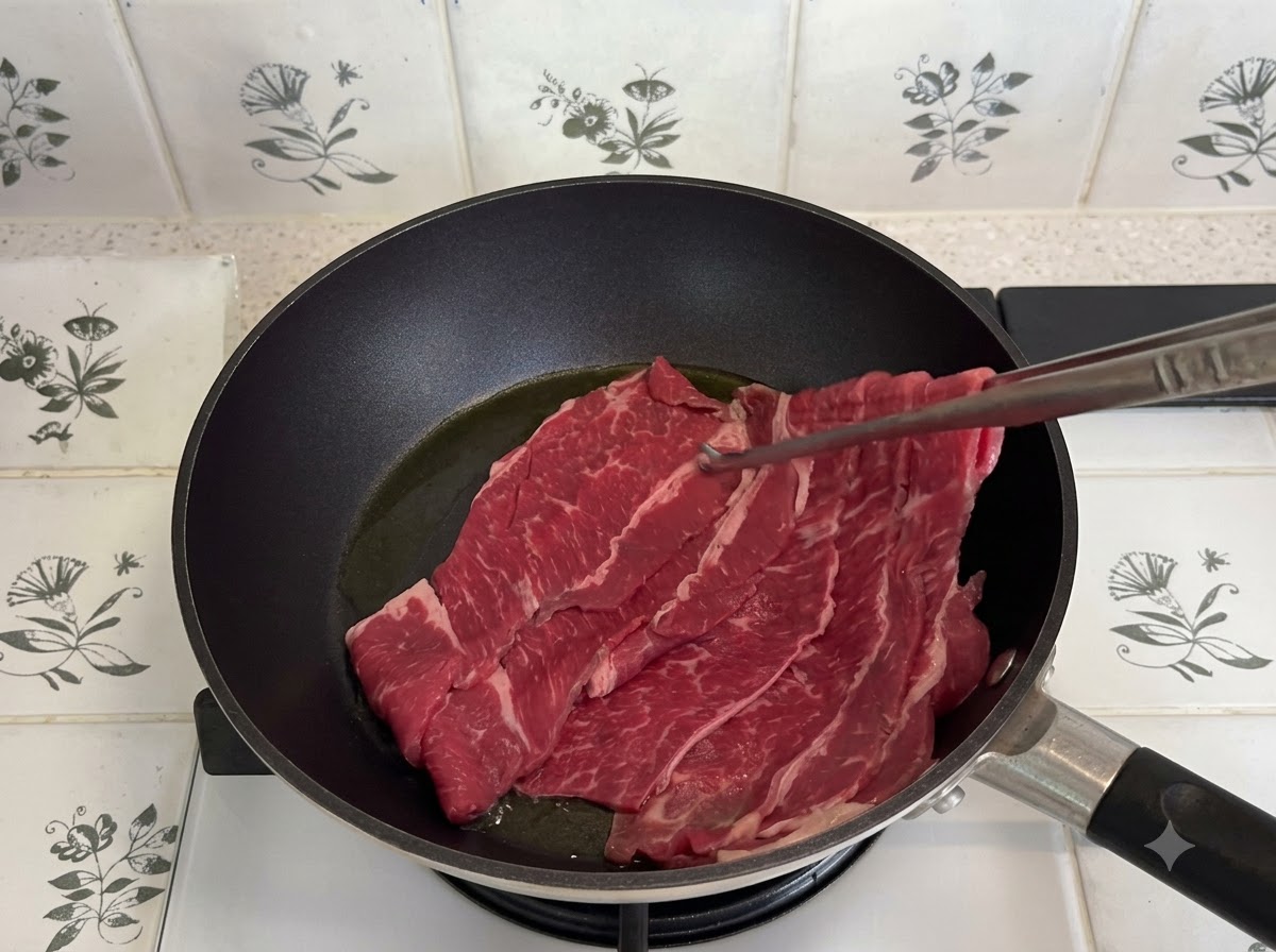 Thinly sliced fatty beef being pan-fried in a dark skillet with metal tongs.