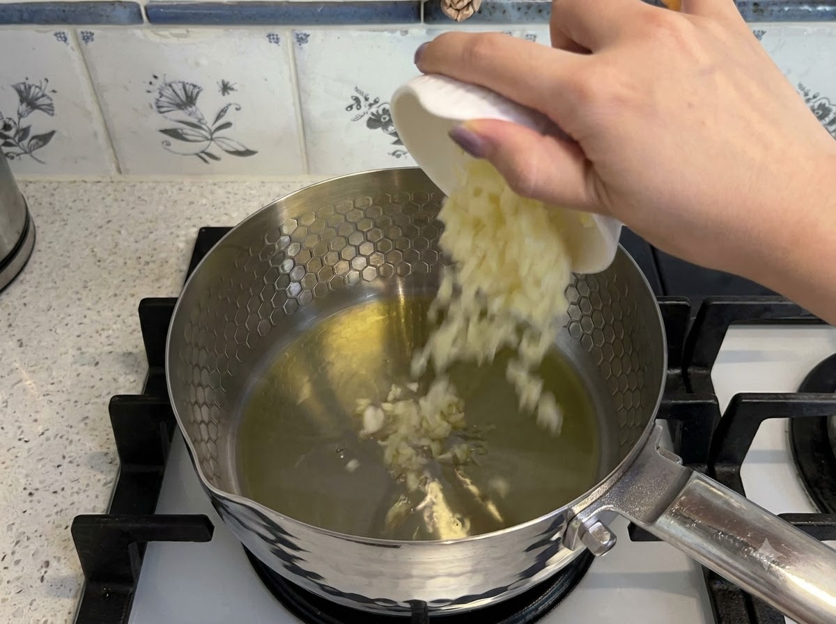 Minced garlic and diced tomatoes being added to hot oil in a pot to sauté.