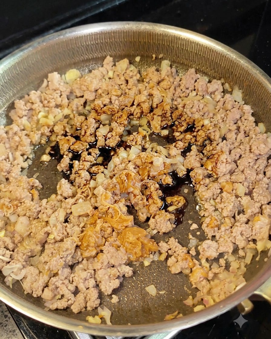 Dark soy sauce being poured from a small spoon onto minced meat and onions in a frying pan.