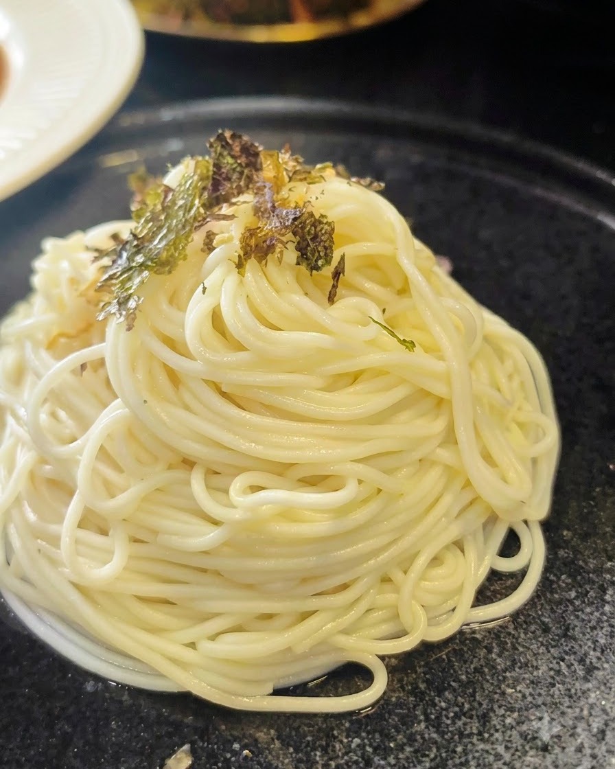 Crushed dried seaweed flakes being sprinkled over a neat spiral mound of cooked ramen noodles on a dark plate.