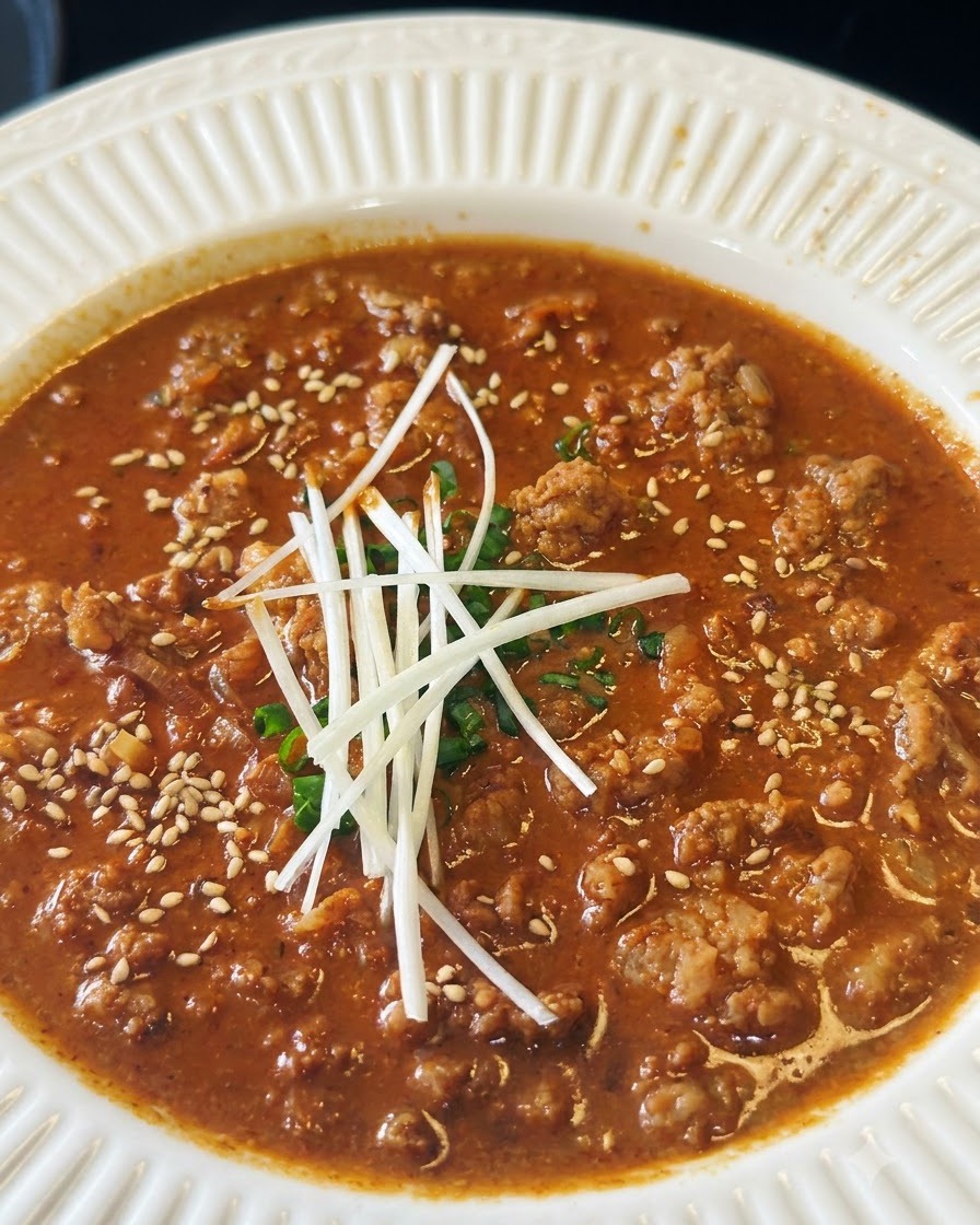 Toasted white sesame seeds being sprinkled across a bowl of savory meat sauce garnished with green onions and scallion strips.