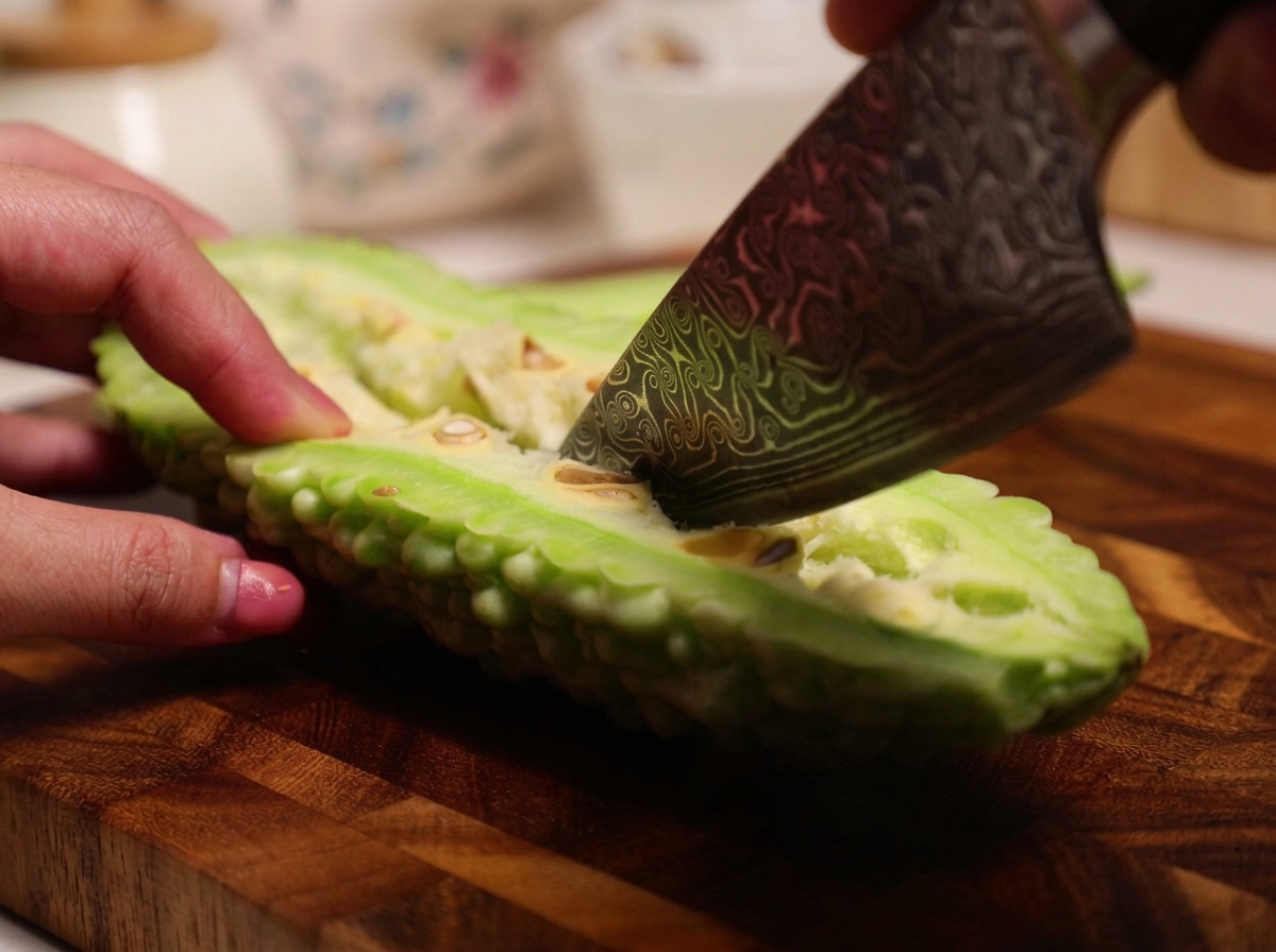 A knife being used to scrape the spongy white pith and seeds from the inside of a halved raw bitter melon.
