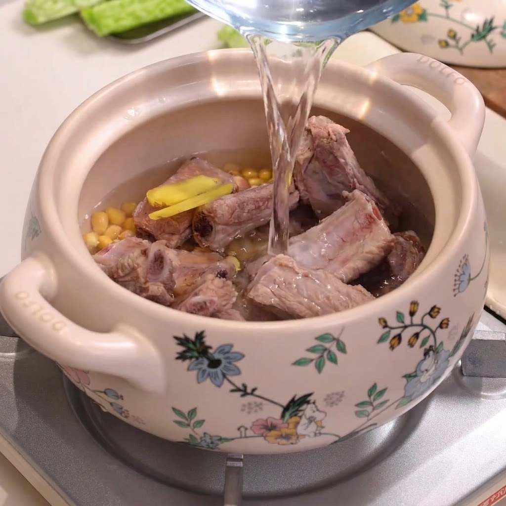Water being poured from a pitcher into a decorated ceramic stew pot filled with pork ribs and soybeans.