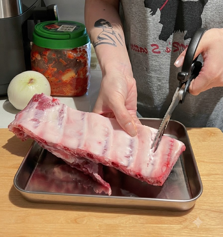 Hands using kitchen scissors to cut a large rack of raw pork ribs into individual pieces on a wooden cutting board.