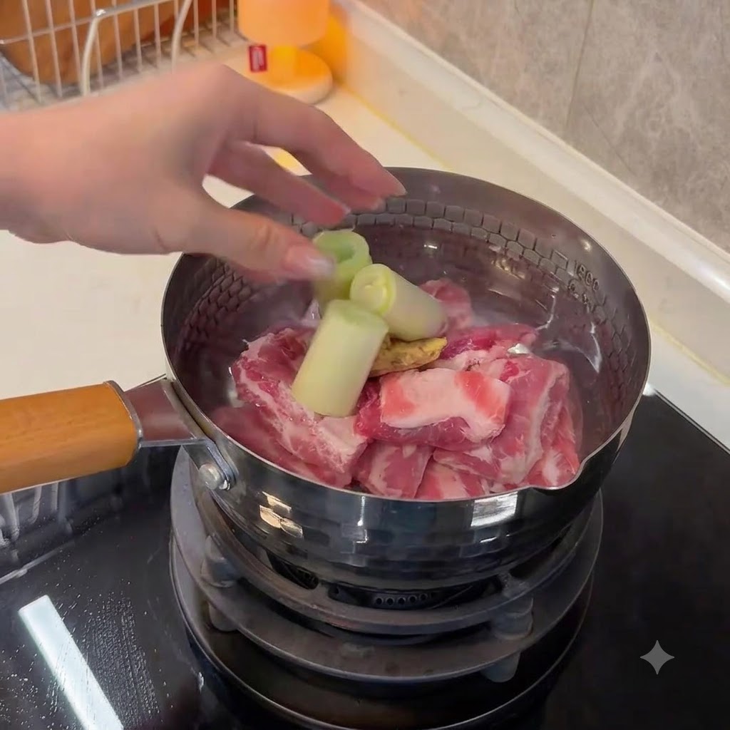 A hand dropping large pieces of green onion and sliced ginger into a pot containing pork ribs and water.
