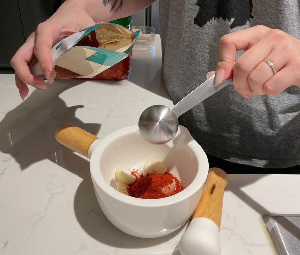 Measuring red chili powder into a white mortar filled with garlic cloves.