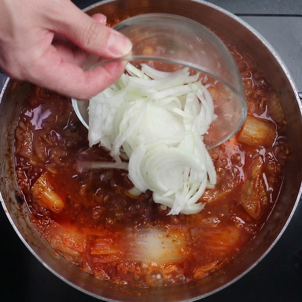 Sliced white onions being poured from a glass bowl into a pot of vigorously bubbling red, spicy kimchi broth.