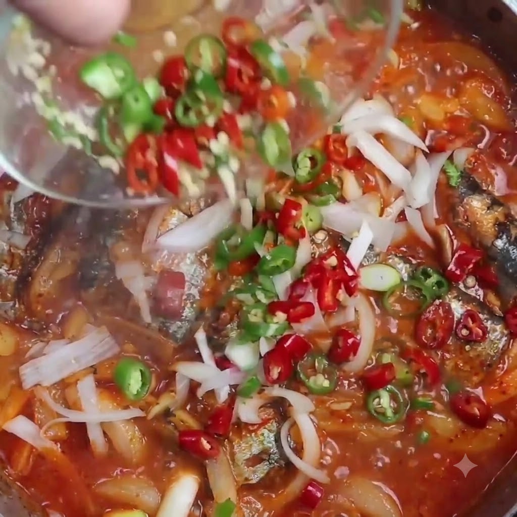 Sliced red and green chili peppers being poured from a transparent small bowl into a simmering pot of braised saury.