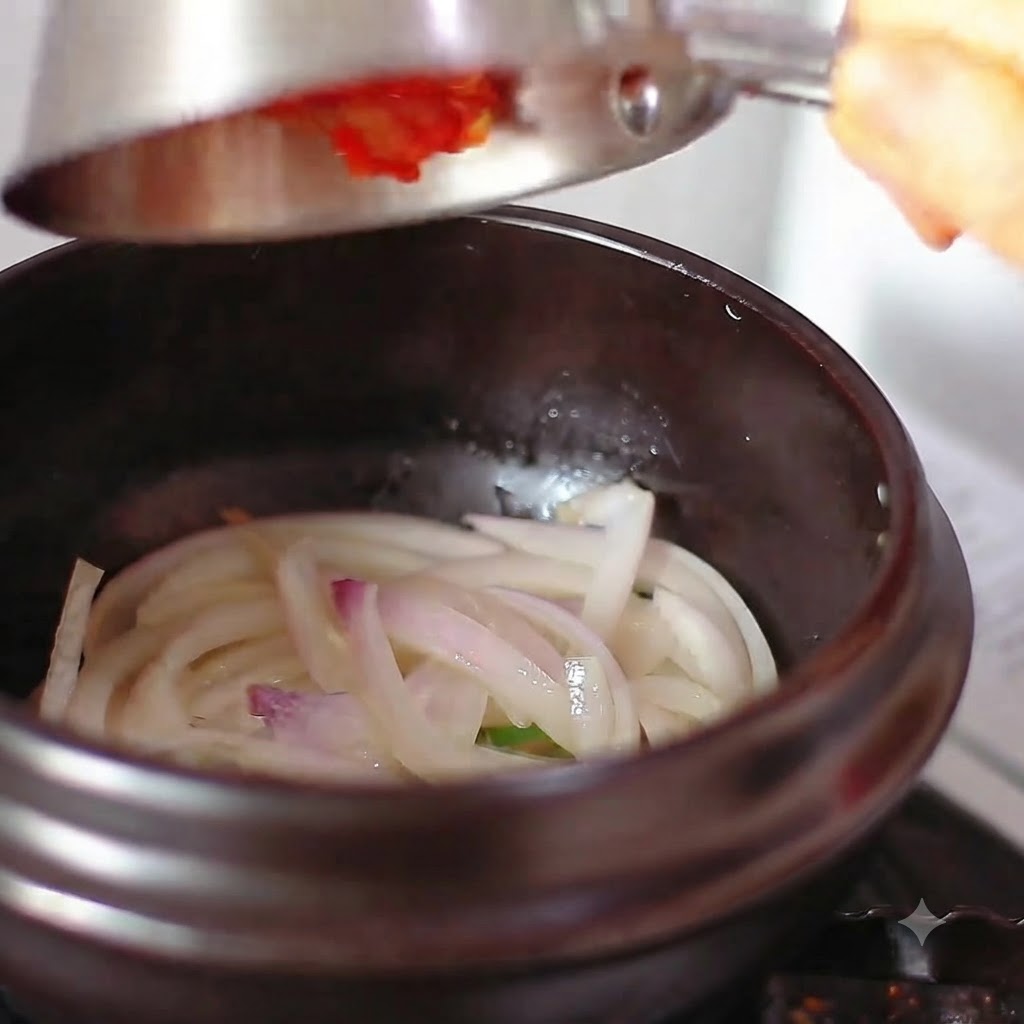 Red chili powder and kimchi being tipped from a metal container into the pot with onions.