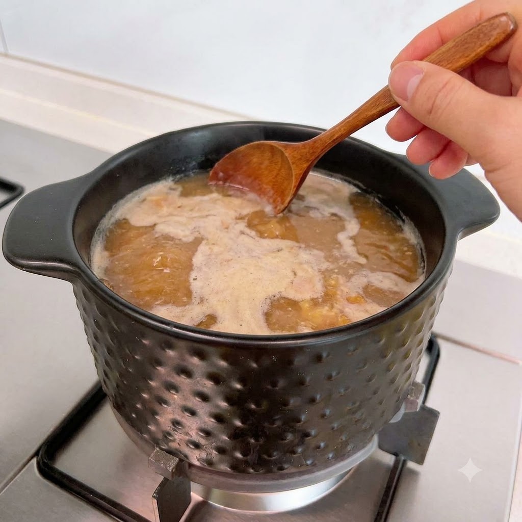 A wooden spoon skimming foam from the surface of simmering soup in a pot.