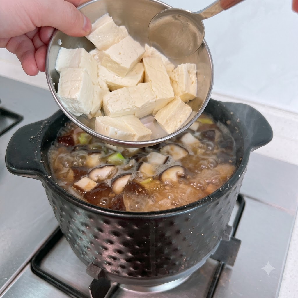 Sliced white tofu being added from a metal bowl into the bubbling stew.