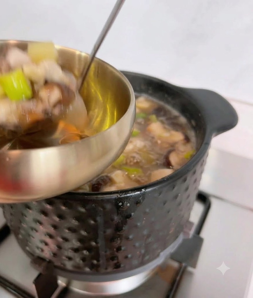 A ladle scooping hot soybean paste soup and ingredients from a black pot into a metallic serving bowl.