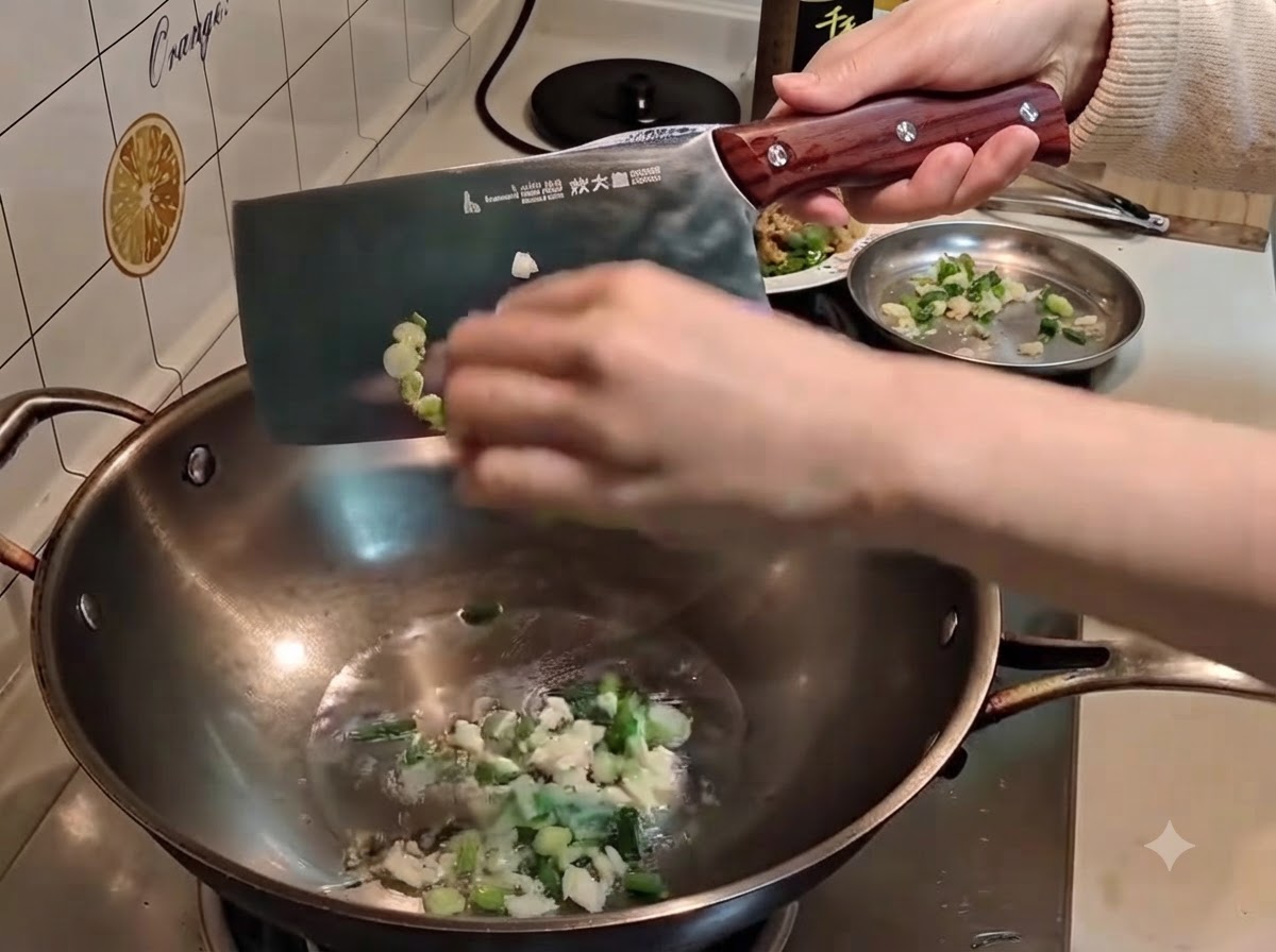 A hand using a large metal cleaver to push chopped scallions and garlic into hot oil in a wok.