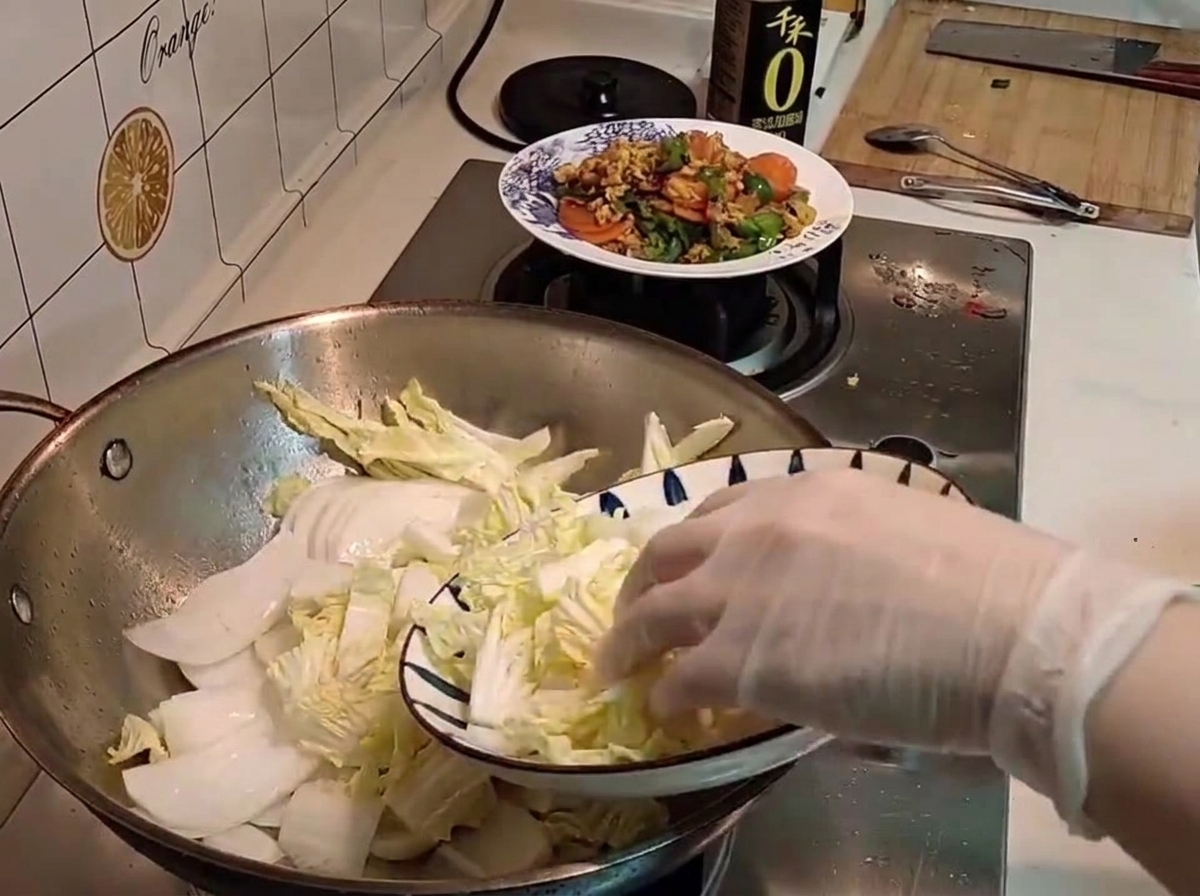 A gloved hand transferring freshly sliced raw napa cabbage from a patterned plate into a wok.