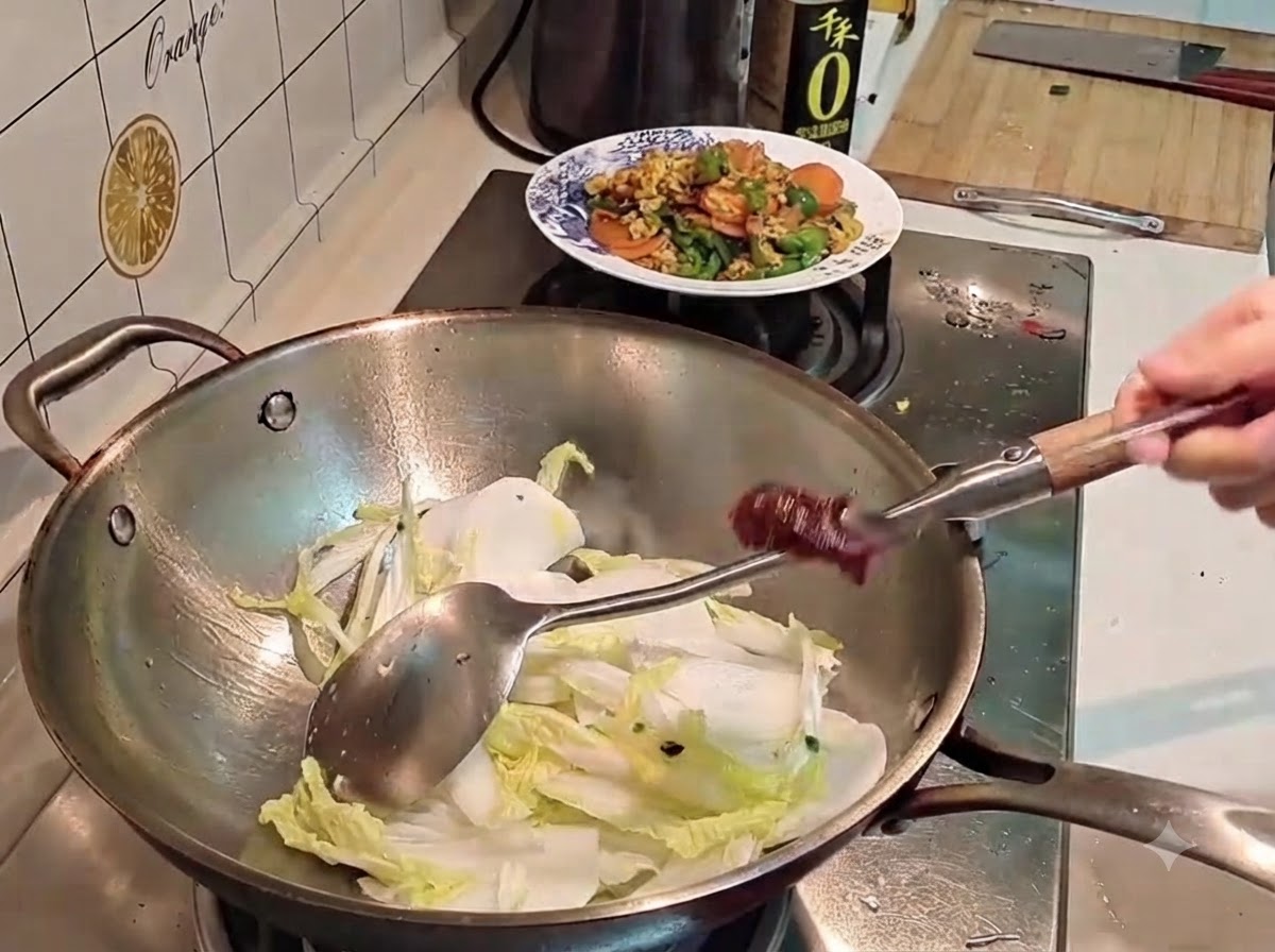 A hand holding a metal spoon loaded with dark red Korean chili paste over a wok filled with cabbage and radish.