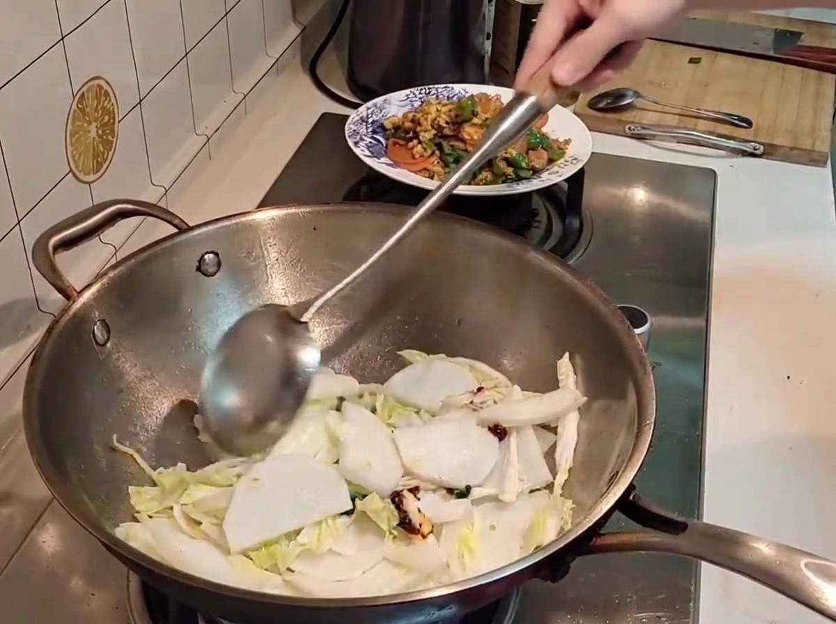 A metal spatula tossing pieces of napa cabbage and white radish with red chili paste inside a hot wok.