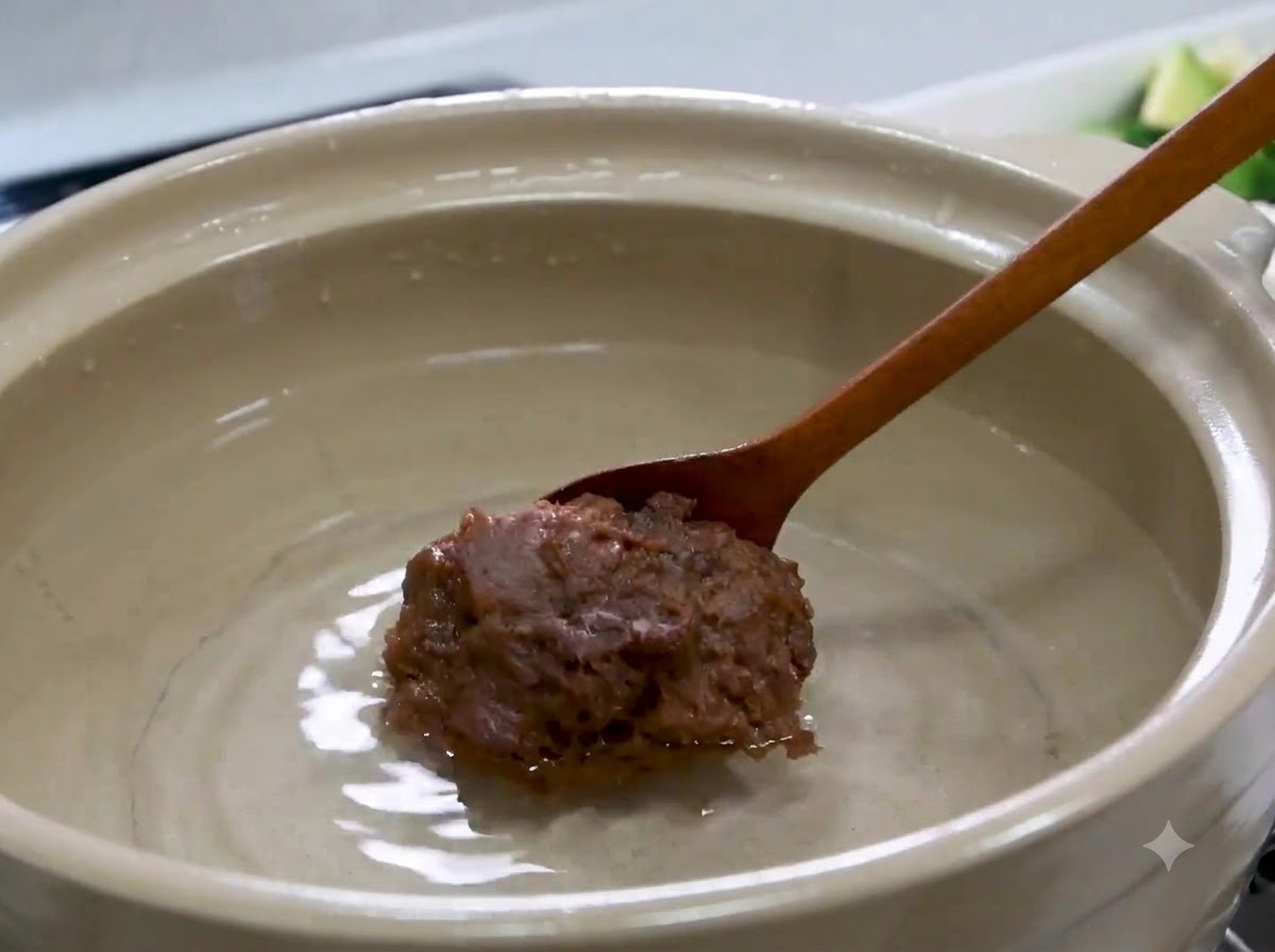 A wooden spoon holding a large lump of Korean soybean paste over the water inside a pot.
