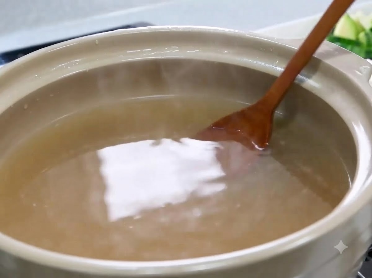 A wooden spoon stirring and dissolving soybean paste inside a pot of light brown broth.