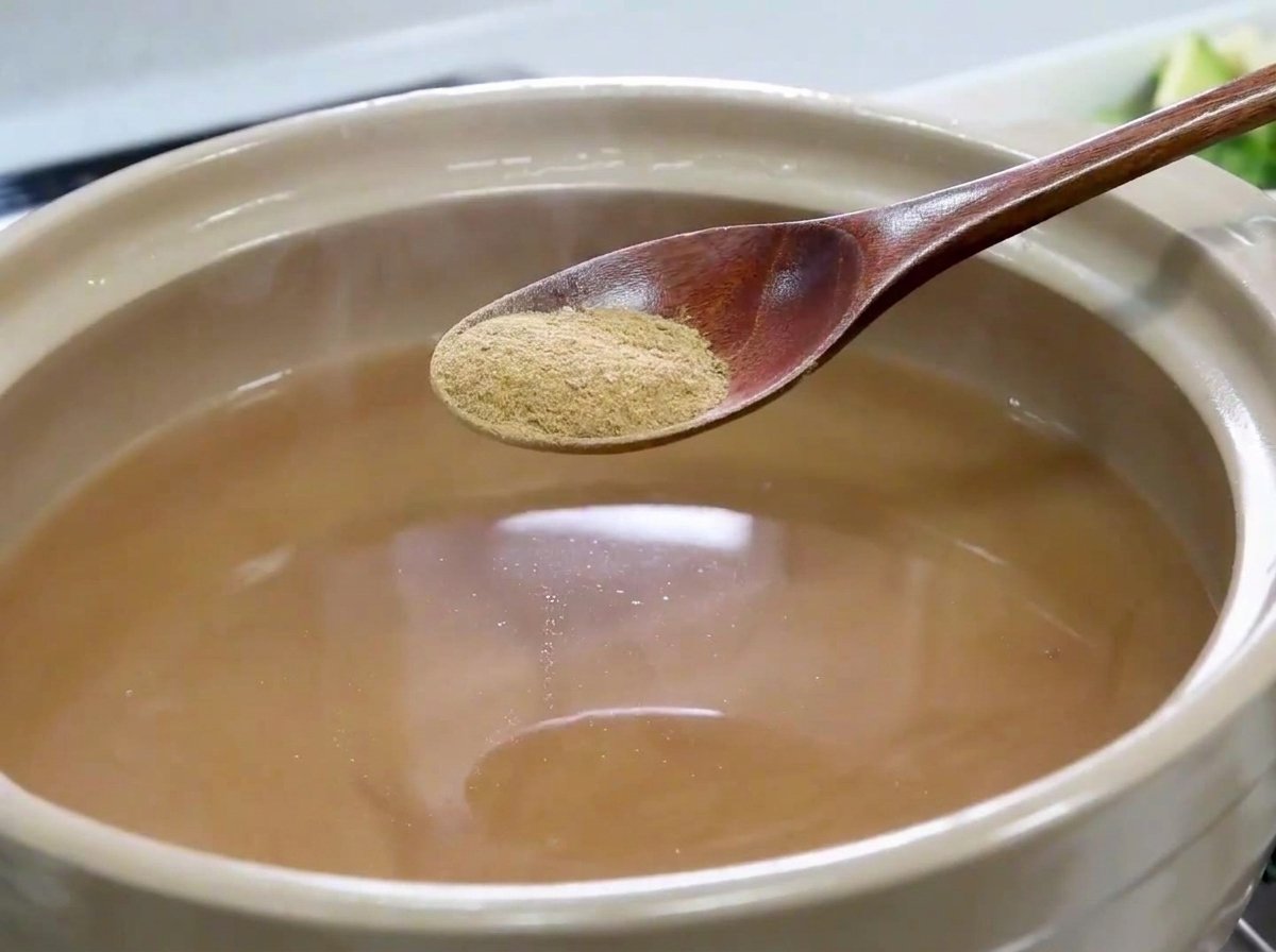 A wooden spoon holding fine anchovy powder above a pot filled with light brown broth.