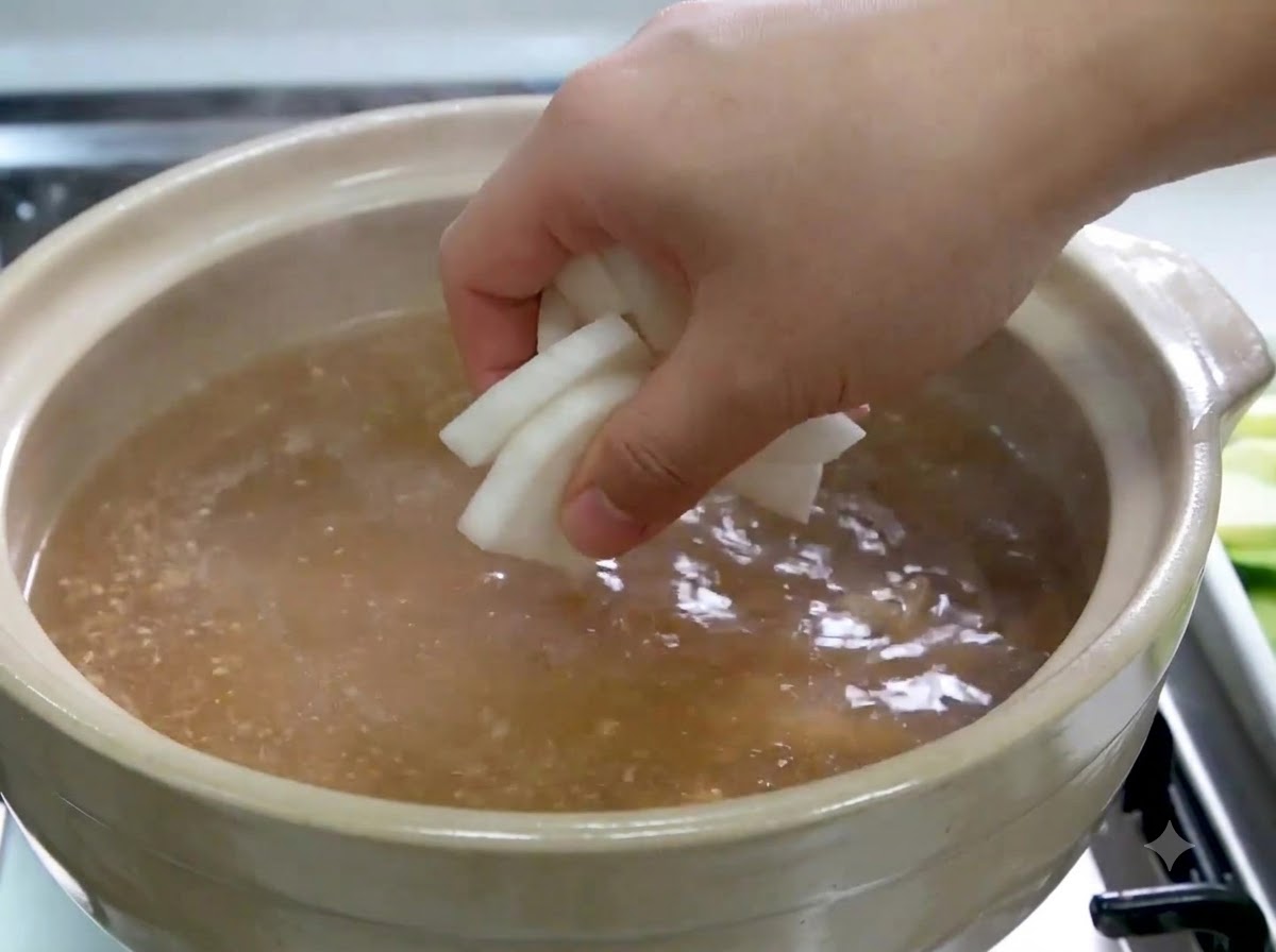 A hand placing white radish slices into a simmering pot of broth.