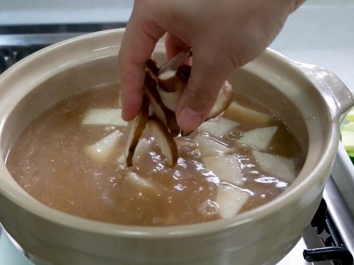 A hand dropping sliced shiitake mushrooms into a pot containing broth and radish slices.