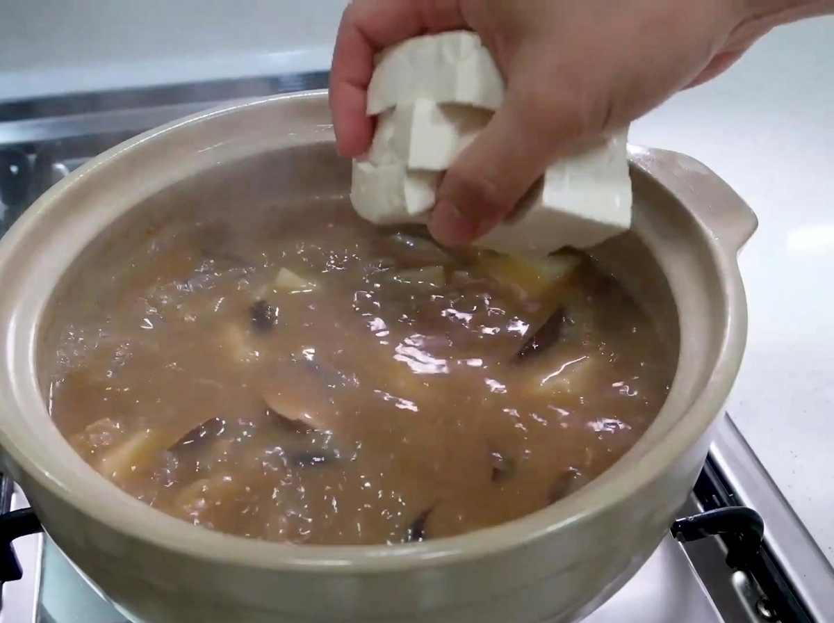 A hand carefully dropping fresh, white tofu cubes into a pot of bubbling brown stew.