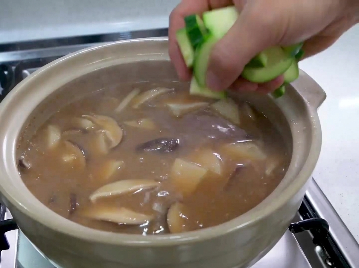 A hand dropping chopped pieces of raw green zucchini into a bubbling Korean stew.