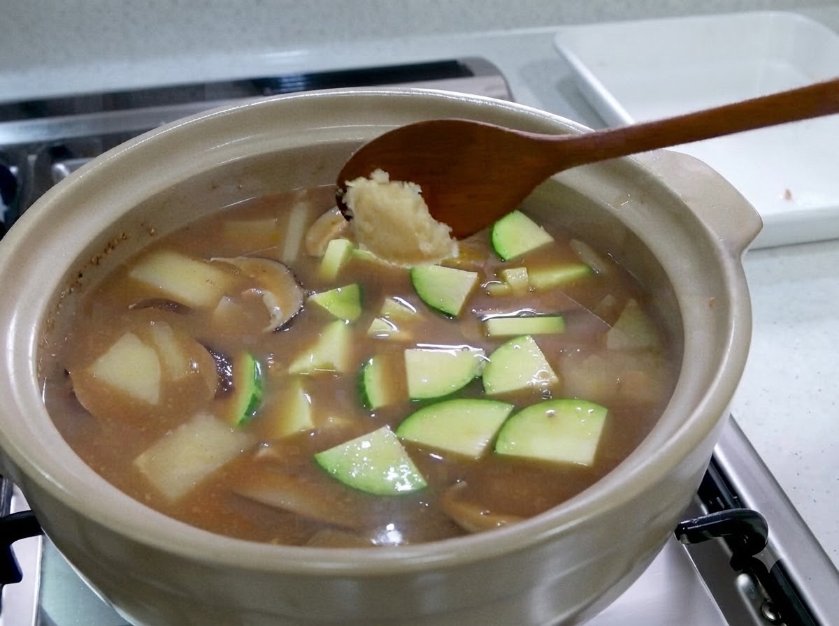 A wooden spoon holding a dollop of minced garlic over a bubbling stew filled with zucchini slices.