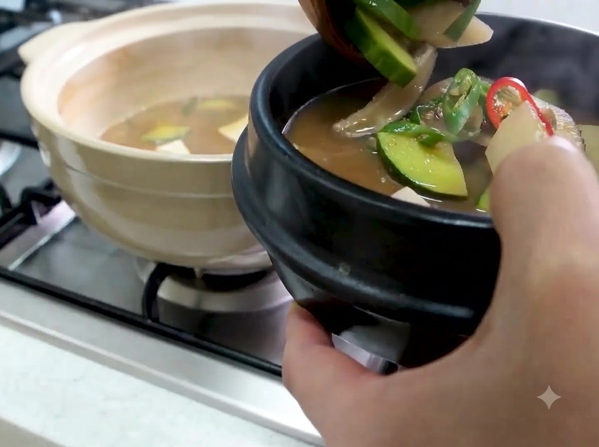 A ladle transferring hot Doenjang Jjigae with zucchini and vegetables into a black stone serving bowl.