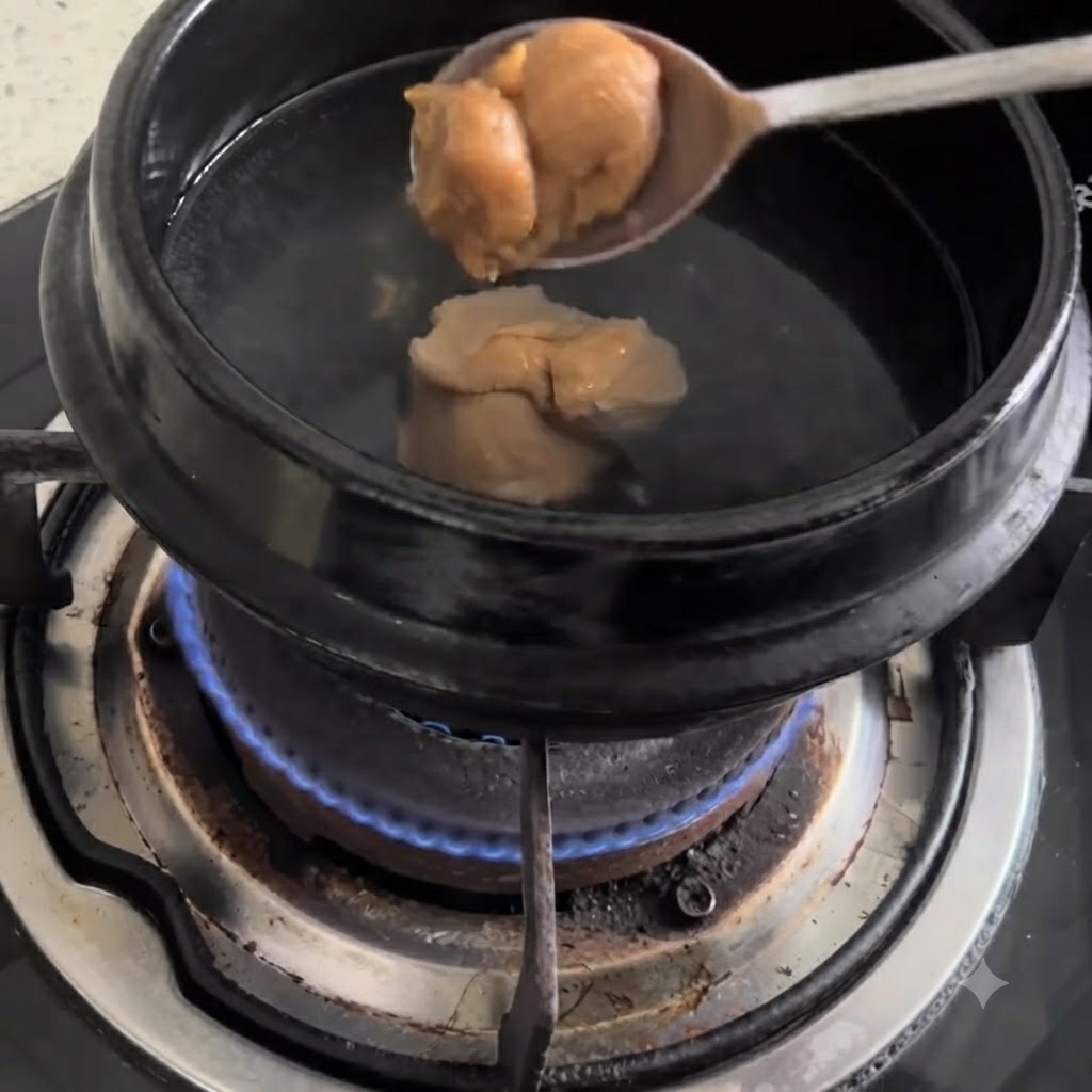 A spoonful of soybean paste being dropped into a black pot of boiling water on a gas stove.