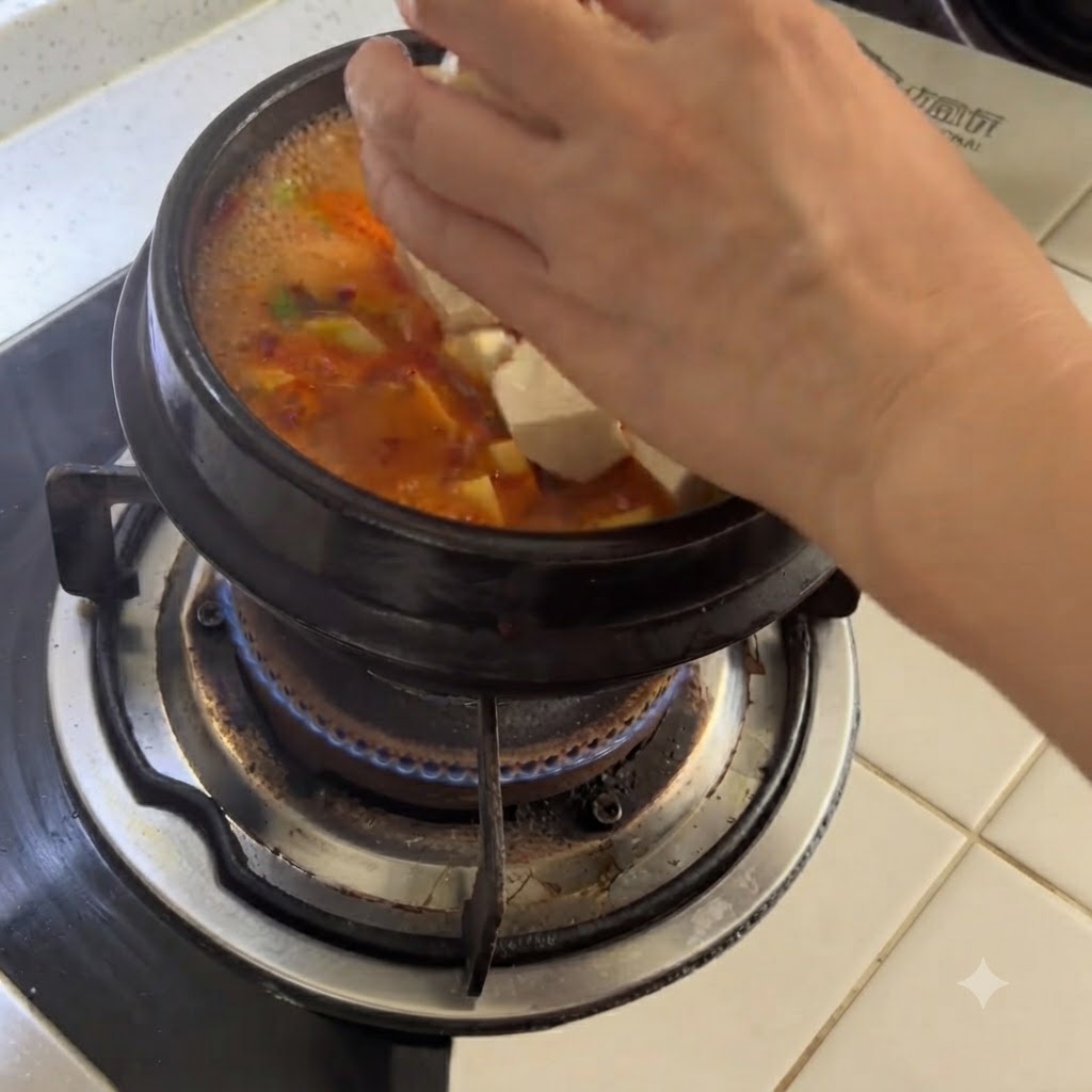 Cubed white tofu being dropped by hand into a simmering clay pot of spicy soup.