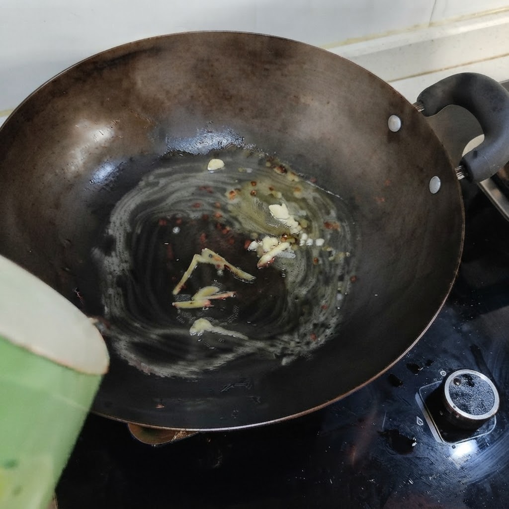 Boiling water being poured from a green container into a wok containing melted lard, ginger, and garlic.