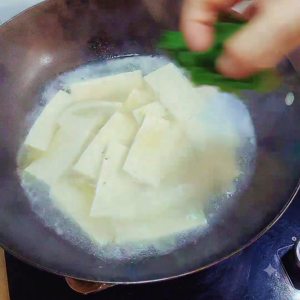 A hand dropping chopped green scallions over the finished tofu soup in the wok.