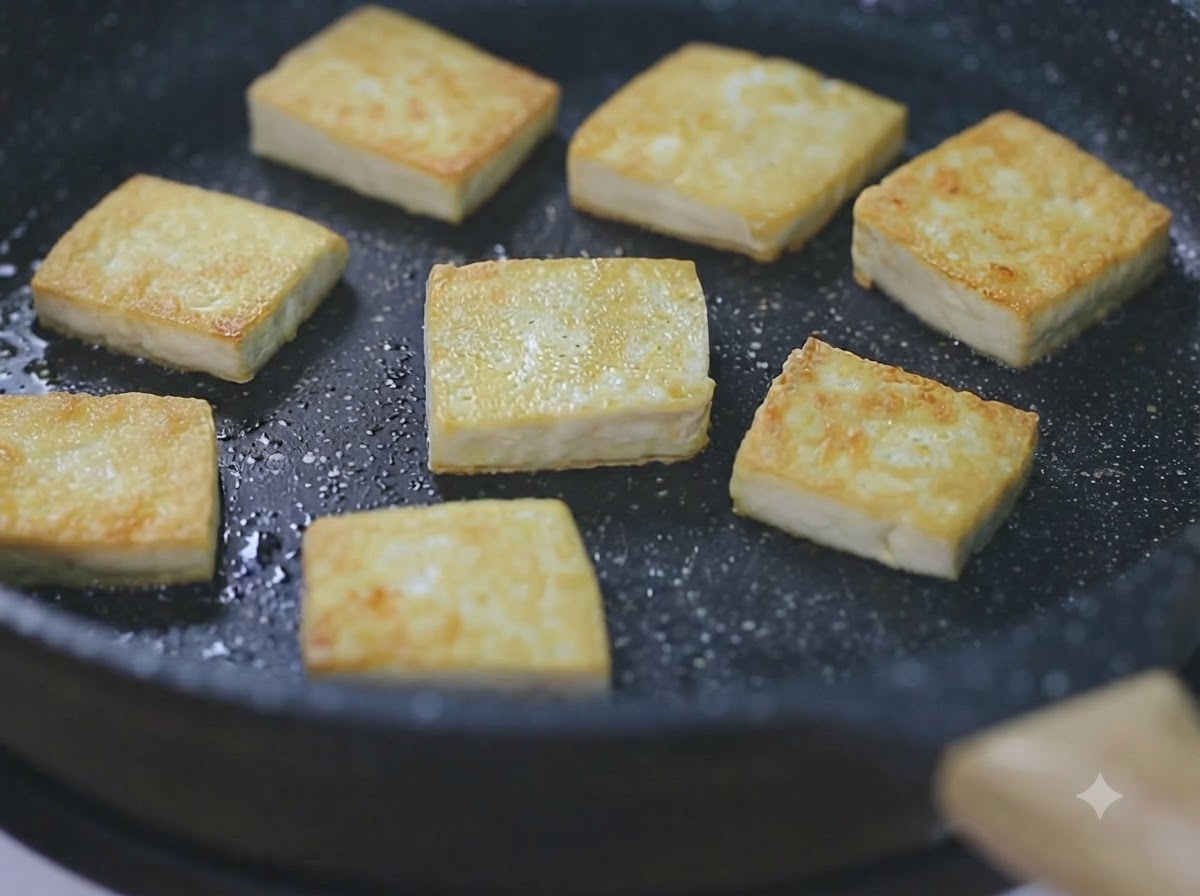 Several golden-brown tofu cubes sizzling in a black skillet over low heat.