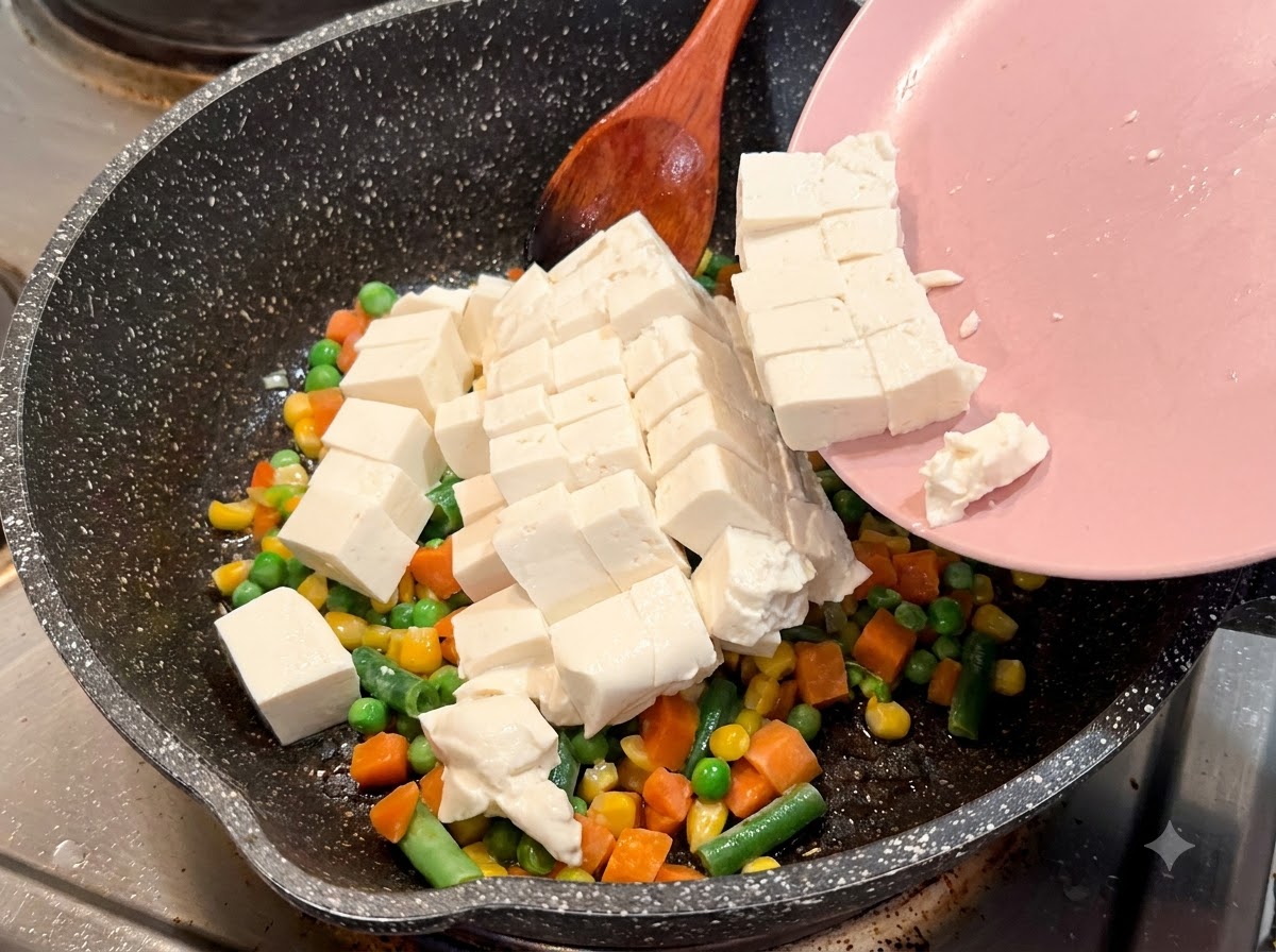 Cubed soft tofu sliding off a pink plate into a speckled frying pan with mixed vegetables.