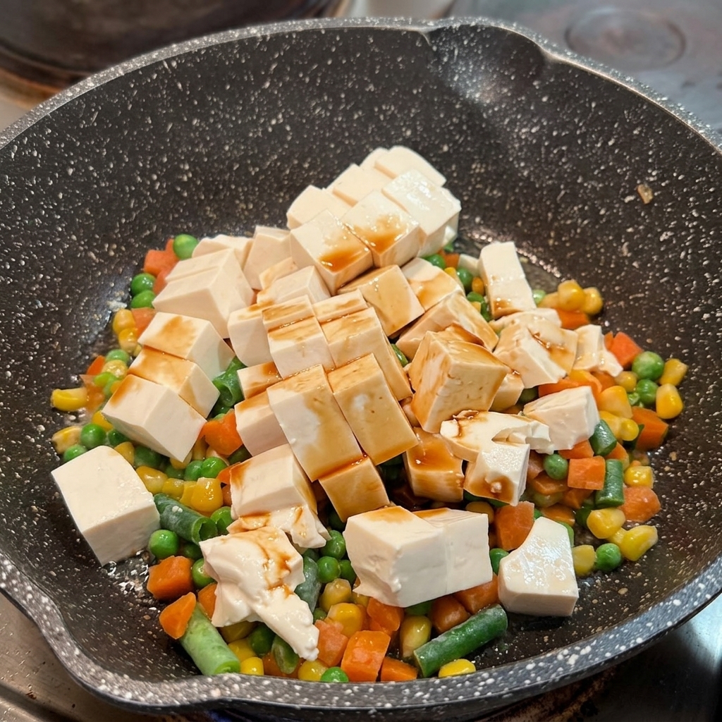 Soy sauce distributed over soft tofu cubes and mixed vegetables in a skillet.