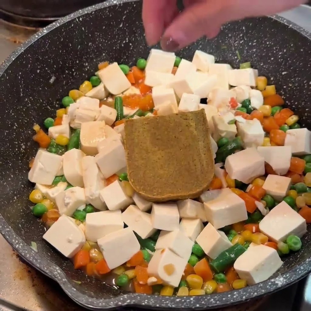A hand placing a block of solid brown curry roux onto tofu and vegetables in a pan.