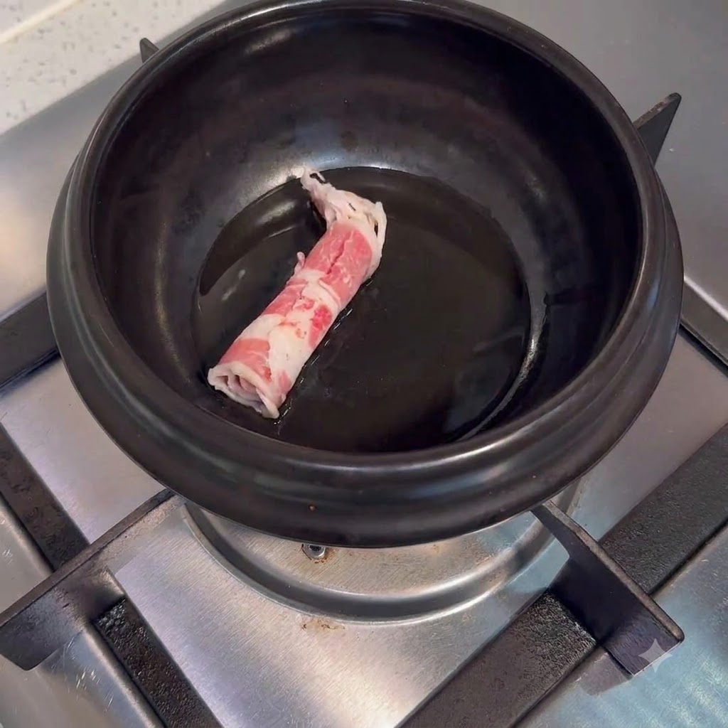 Raw beef slices placed inside a dark ceramic cooking pot with oil on a stove burner.